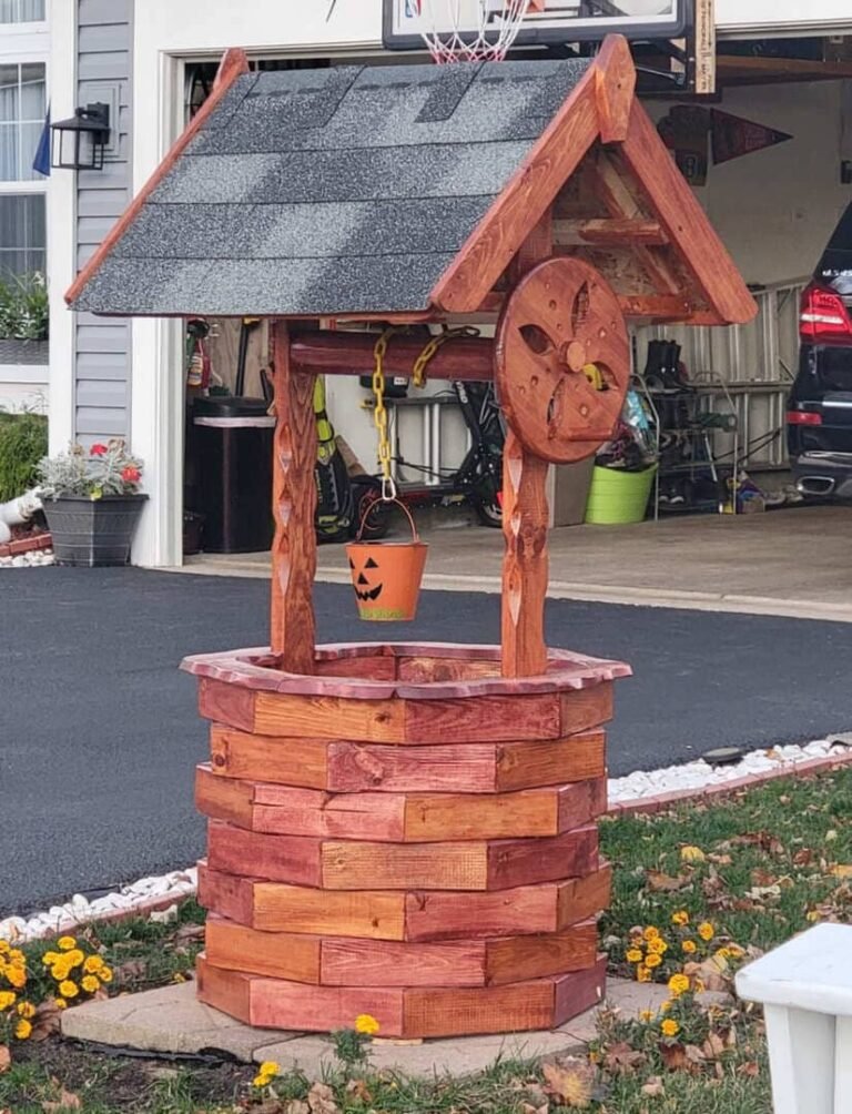 Handcrafted wooden wishing well with a rustic design, featuring a shingled roof, decorative wheel, and a small orange bucket with a jack-o'-lantern face hanging from a yellow chain. The well is placed in a front yard with blooming yellow flowers, near a driveway and an open garage.