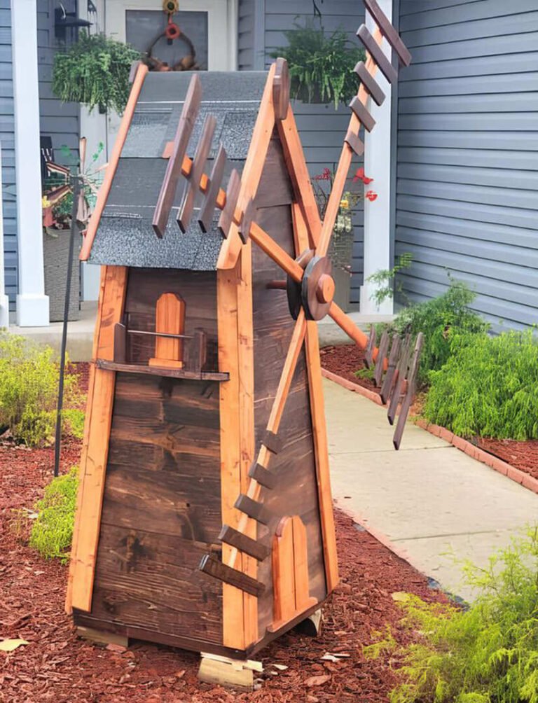 Handcrafted wooden windmill with a rustic design, featuring dark-stained wood panels, a shingled roof, and detailed wooden blades. The structure has small decorative windows and a miniature balcony. Positioned in a landscaped front yard with mulch, greenery, and a walkway leading to a house with a gray exterior and hanging plants.