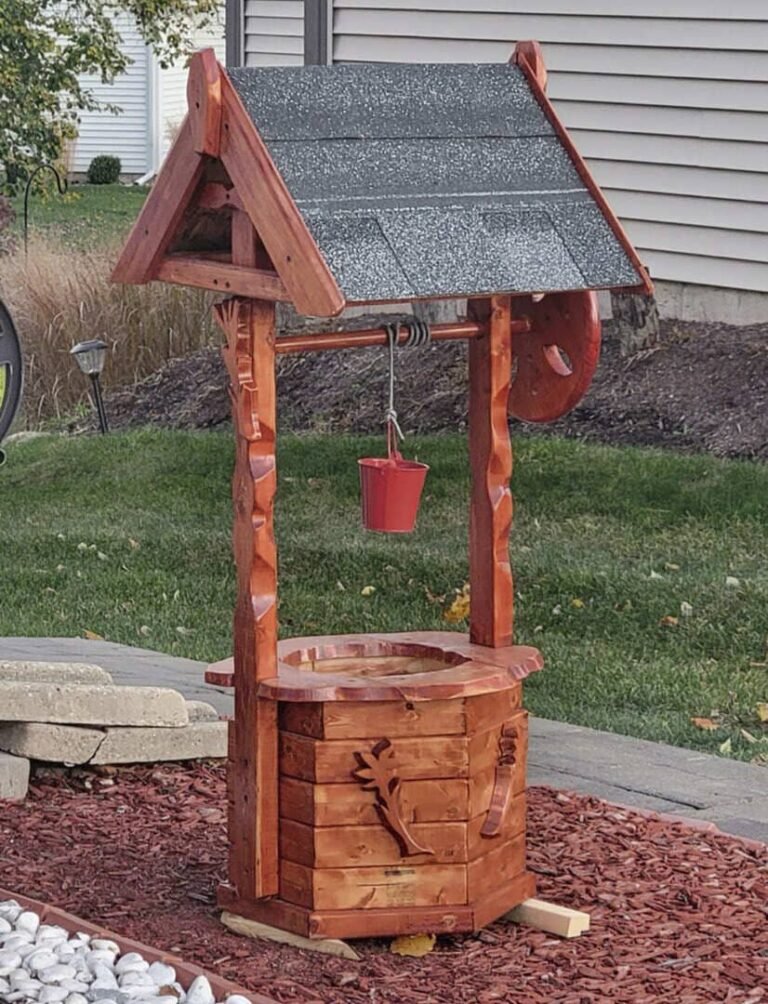 Rustic wooden wishing well with a shingled roof, decorative carved details, and a small red bucket hanging from a metal chain. The well stands on a landscaped area with red mulch, stone edging, and a neatly trimmed lawn, set against a suburban house with beige siding and a garden in the background.