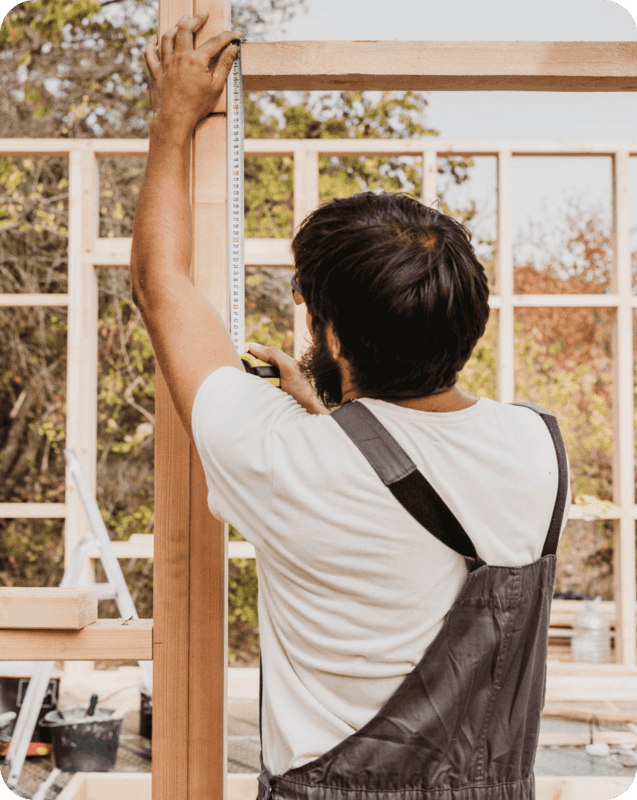 Construction worker in overalls measuring a wooden frame with a tape measure at a building site. The partially built wooden structure is set against an outdoor backdrop with trees and construction tools in the background.