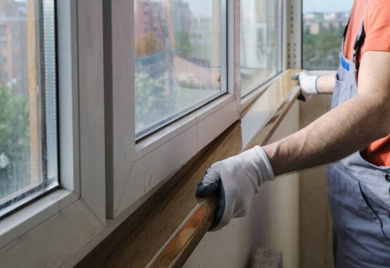 Window repair technician wearing gloves and work overalls installing or repairing a wooden windowsill. The worker is adjusting the sill beneath a double-glazed white-framed window in an apartment with a cityscape view in the background.